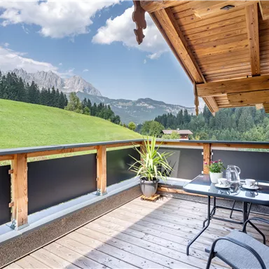 A cozy balcony with a table and chairs. In the background, green hills and mountains can be seen under a blue sky.