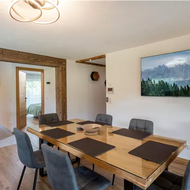 A modern dining room with a large wooden table and six chairs. A picture of mountain landscapes hangs on the wall, while large windows offer a view outside.