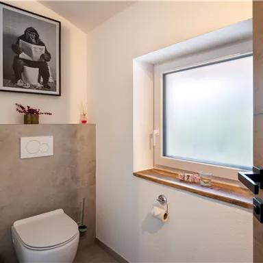 A modern bathroom with a toilet and a window. On the wall hangs a poster-like picture of a person on the toilet.