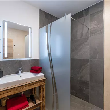 A modern bathroom with a glass shower enclosure and a wooden sink. The walls are decorated with gray tiles, and red towels complete the picture.