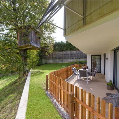 A modern house with a terrace and a cozy seating area. Next to the house is a treehouse, surrounded by lots of greenery.