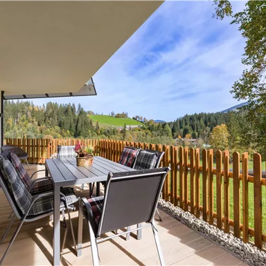 A cozy balcony with a dining table and chairs. In the background, a picturesque landscape with meadows and trees stretches out.