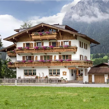 A charming wooden house with balconies and colorful flowers. In the background, mountains and a cloudy sky can be seen.