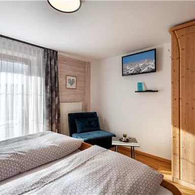A cozy bedroom with a double bed, an armchair, and a small table. A television hangs on the wall and there are large windows with curtains.