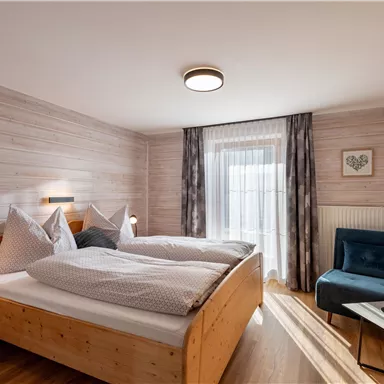A cozy bedroom with two beds and wooden furniture. A blue armchair and a window with curtains provide an inviting view.