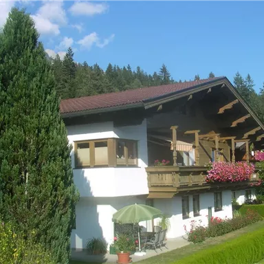 A beautiful house in Bavarian style, surrounded by trees and flowers. On the veranda, there are tables and a sunshade.