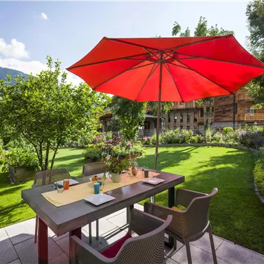 Ein gemütlicher Garten mit einem Tisch und Stühlen unter einem roten Sonnenschirm. Die Umgebung ist grün und blüht, ideal für entspannte Stunden im Freien.