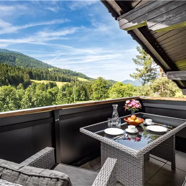 A beautiful balcony with a table and chairs, surrounded by green mountains and trees. The clear sky complements the inviting atmosphere.