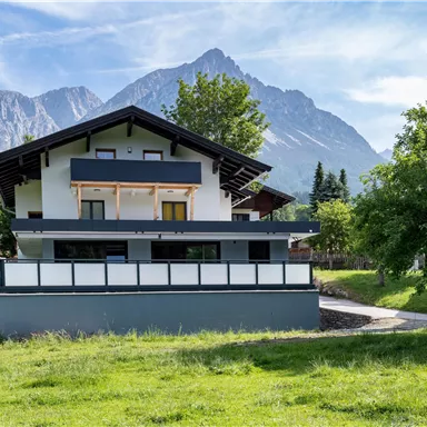 A modern house in front of an impressive mountain landscape. The green meadow and the trees give the scene a tranquil atmosphere.