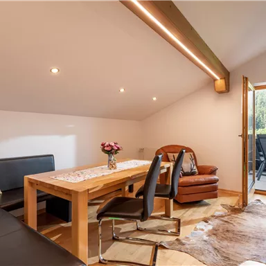 A cozy living space with a dining table, chairs, and a sofa. An open door leads to a balcony outside with a view of nature.