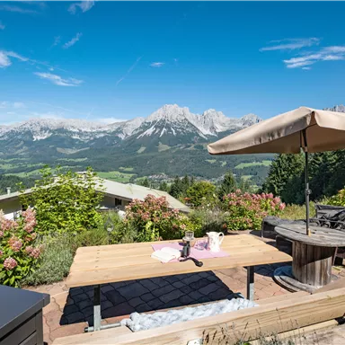 A beautiful terrace with a table and chairs, surrounded by green plants. In the background, impressive mountains are visible under a clear blue sky.