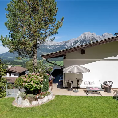 A beautiful house with a garden and terrace, surrounded by mountains and a clear blue sky. The outdoor area is inviting with garden furniture and plants.