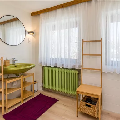 A modern bathroom with a green sink and a large mirror. The windows are adorned with curtains and there is a shelf cabinet with towels.