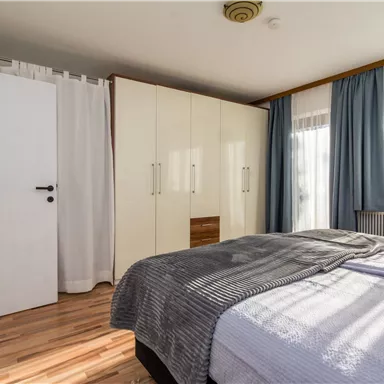 A bright bedroom with a large bed and wooden floor. Curtains and a wardrobe are also visible.