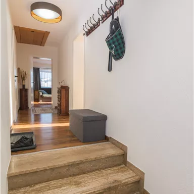A bright hallway with a wooden staircase and modern lighting. Some coat hooks are hanging on the wall, and a gray seating piece is next to the staircase.