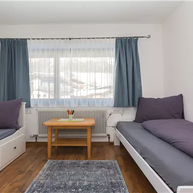 A bright room with two single beds and a small table. Large windows with curtains let in plenty of light.