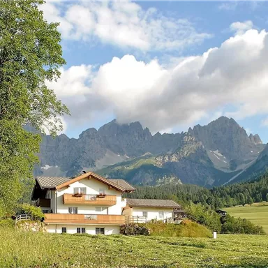 A picturesque house in the midst of nature, surrounded by green meadows and majestic mountains. The sky is partly cloudy, creating a tranquil atmosphere.