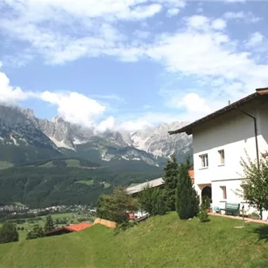 Ein schönes Haus in den Bergen mit einer grünen Wiese. Im Hintergrund sind majestätische Berge und ein klarer Himmel zu sehen.