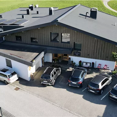 Ein elegantes Holzhaus mit einem großen Dach und bunten Blumen vor der Fassade. Im Vordergrund befinden sich Parkplätze und eine grüne Landschaft im Hintergrund.