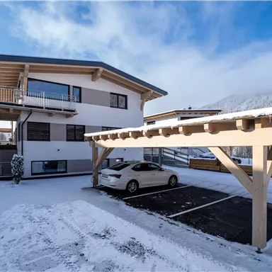 A modern house with a snow-covered parking lot and a covered parking space. In the background, mountains and a blue sky can be seen.