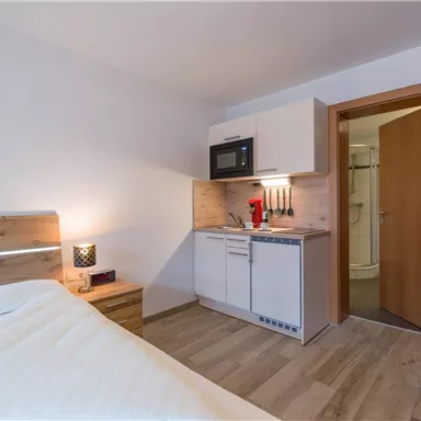 A modern room with a double bed and a small kitchenette. The walls are bright and the floor is wooden.