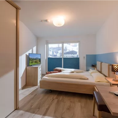 A modern room with two beds, a television, and plenty of natural light. The walls are designed in soft colors and the floor is made of wood.