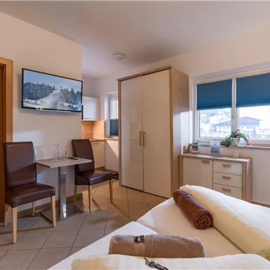 A cozy room with two beds, a table, and two chairs. The windows offer a view of the surroundings, and there is a TV mounted on the wall.