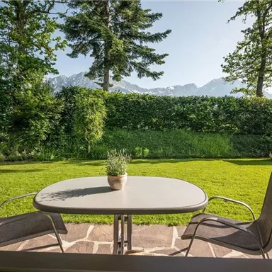 Ein schöner Garten mit einem Tisch und zwei Stühlen. Im Hintergrund sind Bäume und Berge zu sehen.