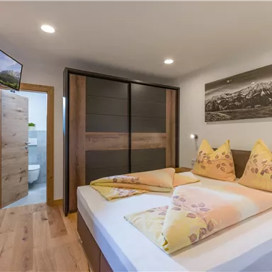 A cozy bedroom with a double bed and colorful pillows. In the background, there is a closed wardrobe and access to the bathroom.