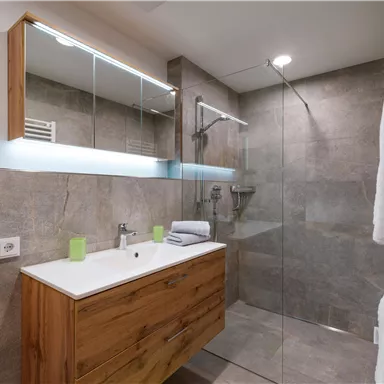A modern bathroom with a glass shower and wooden furniture. There is appealing lighting and fresh towels.