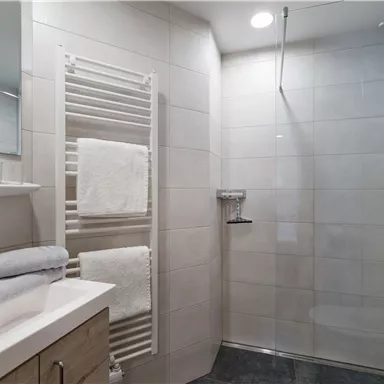 A modern bathroom with a shower and a sink. White tiled floor and a towel rack are also visible.