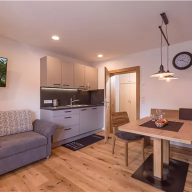 A modern living kitchen with a cozy sofa and a dining table. The decor is bright and inviting, with a television and a clock on the wall.