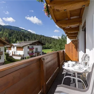 A cozy balcony with chairs and a coffee cup, surrounded by green mountainous terrain. In the background, traditional alpine houses and a clear blue sky can be seen.