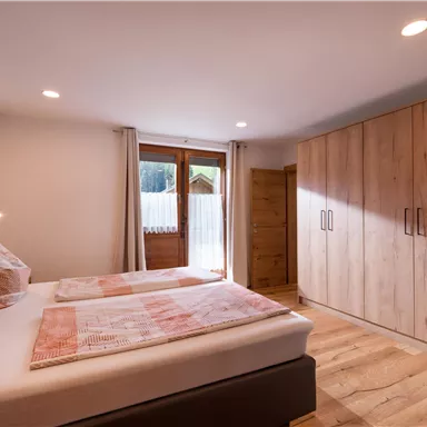 A cozy bedroom with two beds and large windows. The room features warm wooden tones and a wooden wardrobe.