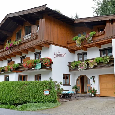 A charming house in alpine style with wooden cladding and colorful flowers. It has several windows and a small garden area.