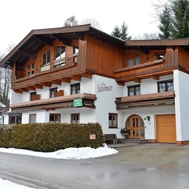 A charming alpine building with wooden cladding and large windows. It is located in a snow-covered environment, surrounded by trees.
