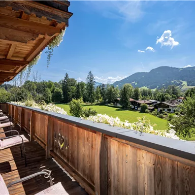 Ein Balkon mit Holzgeländer und gemütlichen Stühlen. Im Hintergrund sieht man eine grüne Wiese und Berge unter einem blauen Himmel.