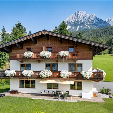 Ein charmantes Haus mit einem Holzverandabereich und blühenden Blumenkästen. Im Hintergrund sind majestätische Berge und eine grüne Wiese zu sehen.