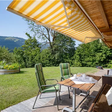 Ein gemütlicher Terrassenbereich mit zwei Stühlen und einem Tisch. Im Hintergrund sieht man eine grüne Wiese und sanfte Hügel.