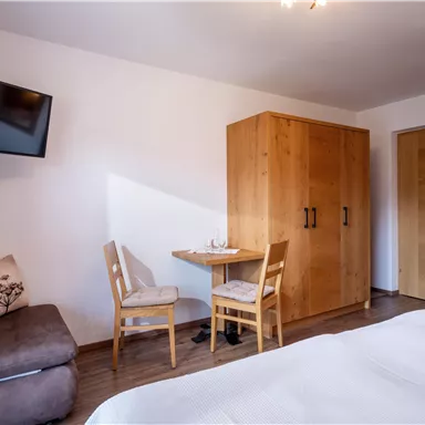Ein gemütliches Zimmer mit einem Bett, einem Tisch und zwei Stühlen. An der Wand hängt ein Fernseher und es gibt einen Kleiderschrank aus Holz.