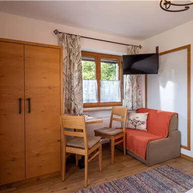 Ein helles Zimmer mit Holzmöbeln und einem kleinen Essbereich. Es gibt einen Fernseher und große Fenster mit Vorhängen.