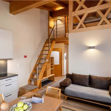 Eine moderne Ferienwohnung mit hellen Wänden und Holzdetails. Die gemütliche Sitzecke und die Treppe führen zu einem Schlafbereich im Obergeschoss.
