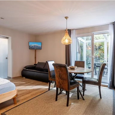 Ein modernes Wohnzimmer mit einem bequemen Sofa, einem Esstisch und einem Fernseher. Große Fenster bieten Zugang zu einem Gartenbereich.