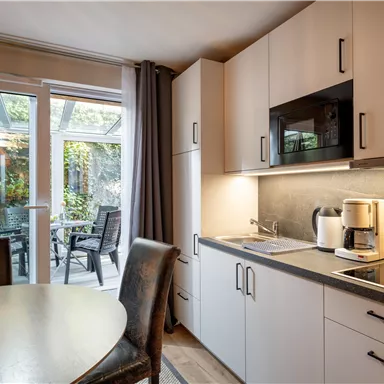 Eine moderne Küche mit hellen Schränken und einem kleinen Esstisch. Durch die Fenster sieht man einen Gartenbereich.