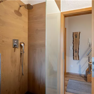Ein modernes Badezimmer mit einer Dusche aus Holz und Glas. Die Tür führt in einen hellen Flur mit Holzdetails.