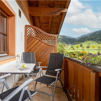 Ein Balkon mit zwei Gartenstühlen und einem Tisch. Im Hintergrund sind grüne Wiesen und Berge zu sehen.