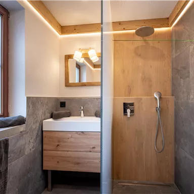 Ein modernes Badezimmer mit einer Dusche und einem Holzwaschbecken. Die Wände sind mit grauen Fliesen gestaltet, und es gibt einen großen Spiegel.