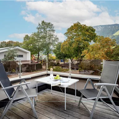 Eine gemütliche Terrasse mit zwei Stühlen und einem kleinen Tisch. Im Hintergrund sind Bäume und Berge sichtbar, unter einem klaren Himmel.