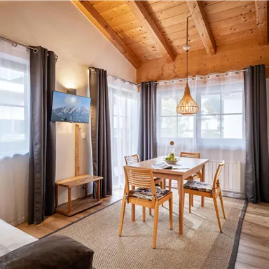 Ein gemütlicher Wohnraum mit Holzdecke und großen Fenstern. Ein Esstisch mit Stühlen ist zentral platziert, und es gibt ein modernes Küchenfenster.