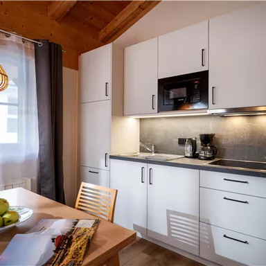 Eine moderne Küche mit weißen Möbeln und Holzdetails. Auf dem Tisch liegen Äpfel und eine Zuckerdose, daneben steht eine Karaffe mit Wasser.